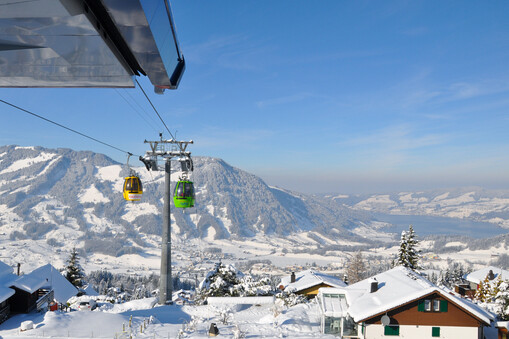 Winterlicher Blick von der Bergstation aus auf den Ägerisee und das ende des Rossberges. In der nähe eines Mastens sind zwei Gondel in gelb und grün zu sehen